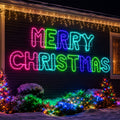 A large LED light sign spelling out 'MERRY CHRISTMAS' in bright multicolor neon-like letters mounted on a dark house exterior at night, with snow on the ground and decorated Christmas trees around it.