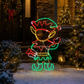 A festive outdoor Christmas decoration shaped like a reindeer or elf, outlined with multicolored LED lights in red, green, yellow, and white, displayed in a snowy front yard at night with decorated trees and a house in the background.