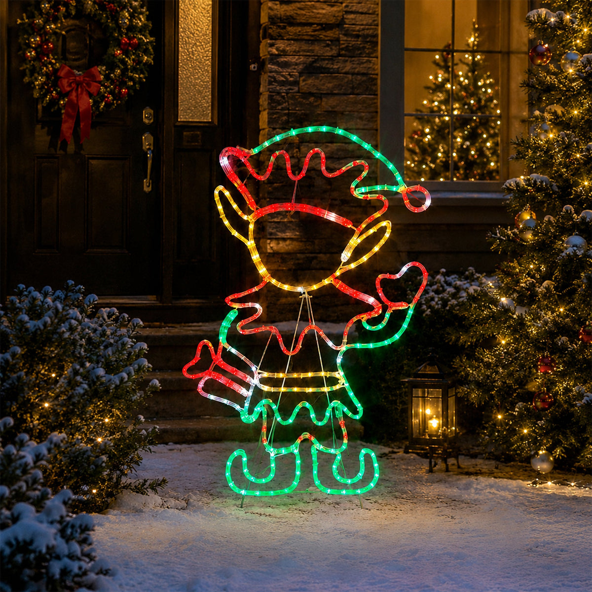 A festive outdoor Christmas decoration shaped like a reindeer or elf, outlined with multicolored LED lights in red, green, yellow, and white, displayed in a snowy front yard at night with decorated trees and a house in the background.